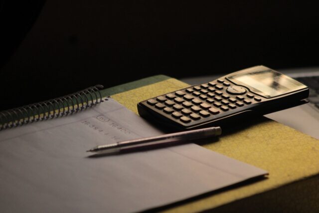 Calculator de birou, un caiet cu calcule și un pix pe o masă de lucru, în timpul stabilirii unei liste de prețuri