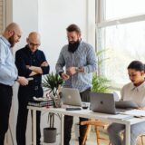 Echipă mică ce lucrează împreună într-un birou luminos, discutând în jurul unui laptop